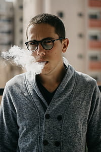 man with black eyeglasses and gray top blowing smoke from his mouth at daytime thumbnail