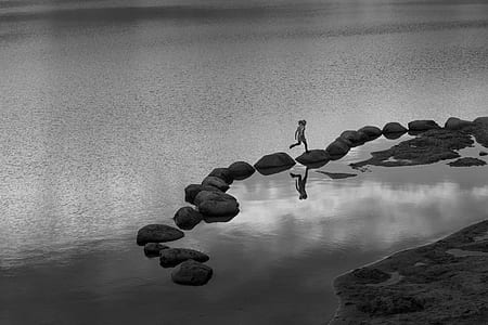 grayscale photography of person hopping on stones in body of water thumbnail