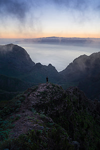 man in black t-shirt and black shorts standing near mountain beside body of water during day time thumbnail