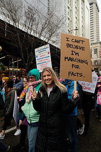 woman wearing black zip-up coat holding brown signage walking in the street thumbnail