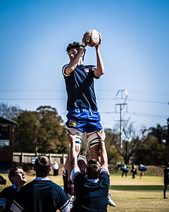 Two Men Carrying Standing Man Holding Rugby Ball over Field With People Across Electric Post Under Blue Sky thumbnail