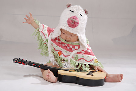 baby wearing knitted cap playing with guitar while sitting on white surface thumbnail