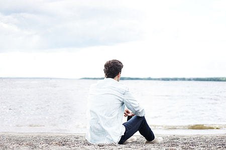 man in white dress shirt and black denim jeans sitting on sand watching seawave during daytime thumbnail