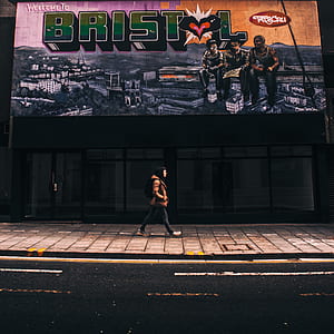 person wearing brown and black hoodie and pants walking near glass panel with Welcome to Bristol graffiti at daytime thumbnail