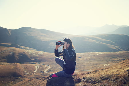 woman sitting on rock formation while using binoculars under white sky thumbnail