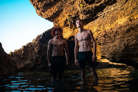 two topless men standing near rock formation with submerged feet during daytime thumbnail
