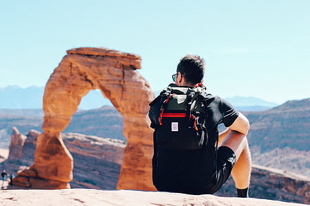 man wearing hydropack backpack sitting on hill facing Delicate Arch in Utah thumbnail