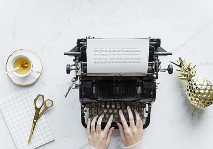 person typing on black typewriter with brown scissors beside and white cup thumbnail