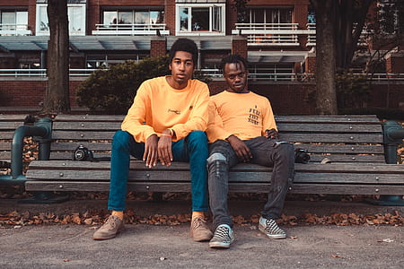 two men sitting on beige wooden bench over brown high-rise building during daytime thumbnail