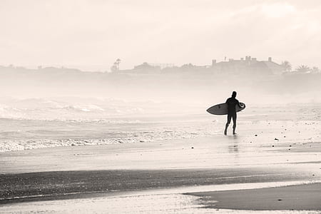 grayscale photo of man holding surfboard in seashore thumbnail