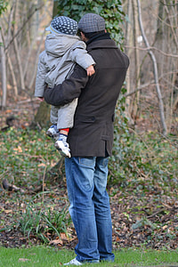 man in black coat carrying baby in grey hoodie jacket white standing on green grass field thumbnail