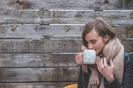 woman in black jacket and brown scarf drinking in white ceramic mug near brown wooden wall during daytime thumbnail
