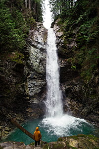 person wearing orange hooded jacket standing in front of waterfall during daytime thumbnail