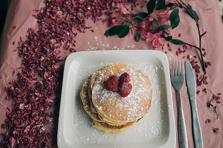 pancake with three strawberries on top of plate beside gray fork and butter knife thumbnail