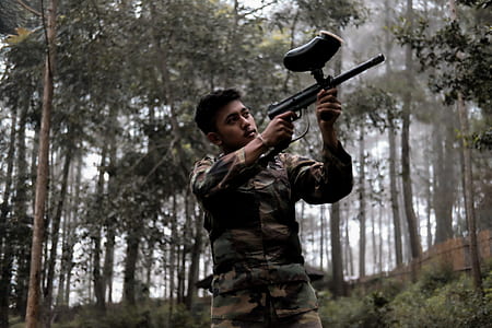 man holding black paintball gun near trees thumbnail