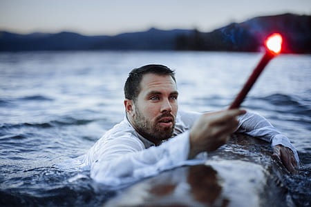 man floating on body of water holding on log with torch during daytime thumbnail