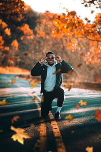 man wearing gray pullover hoodie and black zip-up jacket kneeling on gray concrete road top surrounded with maple trees during daytime thumbnail