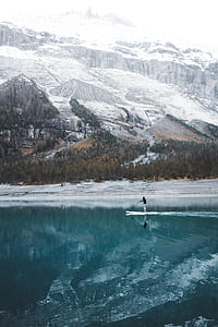 person standing on paddle boat on body of water near snow-covered mountains during daytime thumbnail