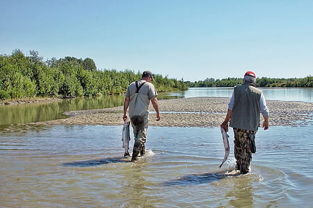two men holding fish while walking on grey body of water near trees during daytime thumbnail