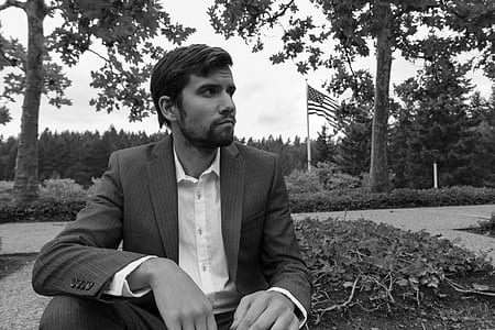 grayscale photo of man sitting beside plant near trees thumbnail