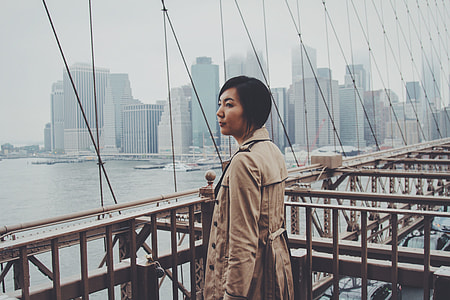 woman wearing brown coat standing on metal bridge with cars running below during daytime thumbnail