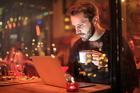 Young White Man With Black Beard Holding Mug in Front of Laptop inside a restaurant thumbnail