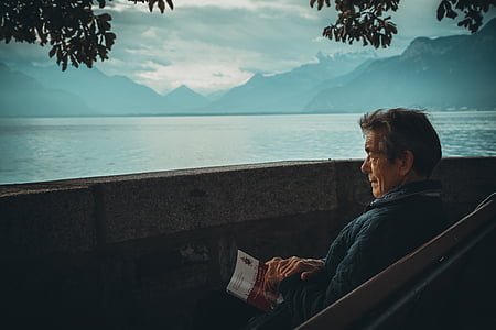 man in blue shirt holding book sitting on chair near body of water thumbnail