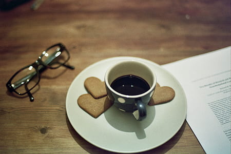 black and white polka-dot ceramic cup filled with black coffee on white saucer near black plastic-frame eyeglasses all on brown wooden table thumbnail