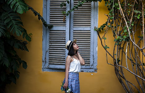 Photo of Woman Wearing White Sleeveless Top and Hat Holding Flowers Near Window thumbnail