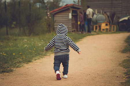 toddler walking on brown soil in front of two men standing near tool shed thumbnail