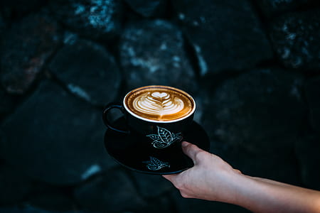 person holding cappuccino filled black ceramic mug and saucer thumbnail