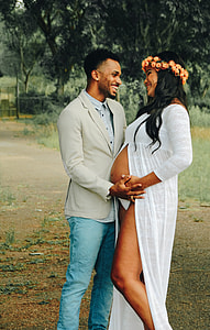 man wearing gray suit jacket and blue jeans standing beside pregnant woman wearing white gown thumbnail
