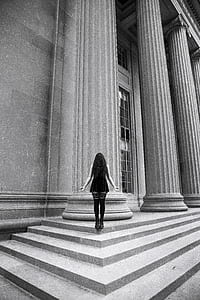 grayscale photography of woman standing between concrete stair and concrete pillar in front of building thumbnail