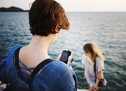 woman holding camera facing woman standing near on water thumbnail