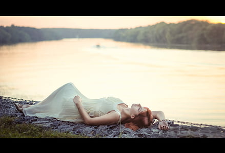 selective focus photography of woman laying on ground near body of water wearing white sleeveless long dress thumbnail