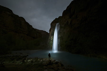 person standing alone on rock facing lighted waterfall during night time thumbnail