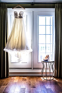 white sweetheart neckline wedding dress hanging in front of window near pair of silver heeled sandals thumbnail