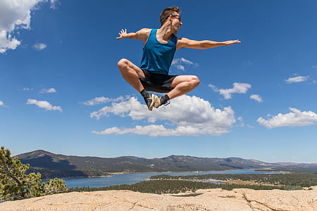 man wearing blue tank top jumping over gray soil near body of water and mountain thumbnail
