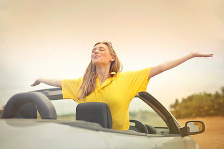 Young Woman In Yellow Shirt Enjoying Weather With Open Arms While Sitting on Convertible Silver Car thumbnail