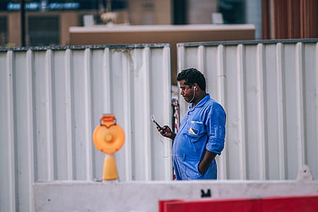 man wearing blue shirt holding smartphone standing near grey corrugated sheet during daytime thumbnail