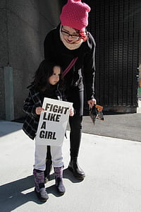 girl in black and white plaid jacket holding white fight like a girl-printed signage during daytime thumbnail