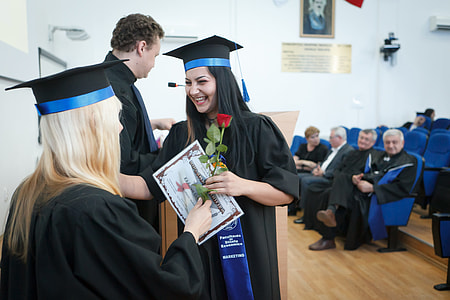 people standing and sitting inside room wearing academic dress and woman holding certificate and red rose smiling thumbnail