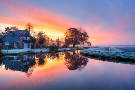 house near river and crop field under orange sky thumbnail