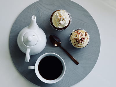white ceramic teapot, cup of coffee, and two cupcakes on grey placemat thumbnail