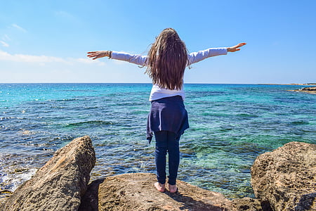 woman wearing white long-sleeved shirt standing on brown stone near ocean with arms wide open during daytime thumbnail