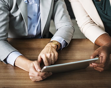 two persons sitting side by side across table and looking at a tablet computer in a well-lit room thumbnail