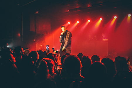 man in black coat standing on stage while singing thumbnail