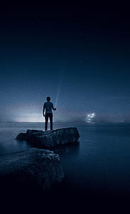man wearing gray shirt and black pants standing on black and gray rock surrounded by water thumbnail