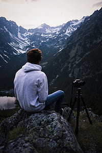 Man in Gray Hoodie Sitting on Gray Stone With Dslr Camera Tripod thumbnail