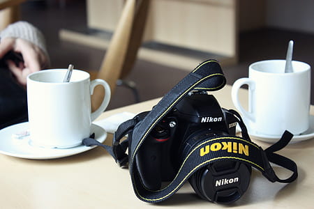 black Nikon DSLR camera beside two white ceramic mugs and saucers thumbnail